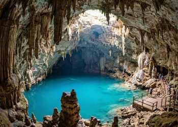 Lago de água azul-turquesa em uma caverna iluminada, cercado por formações rochosas e visitantes em passarela.
