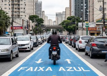 Motociclista segue pela Faixa Azul em corredor sinalizado entre carros na avenida.