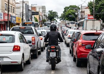 Motociclista passa no corredor com espaço lateral visível entre carros parados.