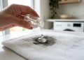 A professional macro photography shot focusing on a brushed stainless steel surface. A hand holding a soft, dark grey microfiber cloth is partially visible, wiping away a dull layer of grease and oily fingerprints. The movement leaves behind a pristine, streak-free path that acts as a mirror, reflecting a blurred high-end modern kitchen with soft pendant lights. The uncleaned area remains matte and textured with smudges. Captured with a Nikon Z7 II, 105mm macro lens, f/5.6, to show the intricate vertical grain of the steel. Lighting: "Clinical Cold Fluorescent" with a sharp side-light to emphasize the metallic luster and the tactile contrast between the greasy and clean zones. No overlaid text. No headlines. No captions. No watermarks.