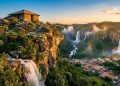Vista panorâmica de cidade histórica cercada por cachoeiras, vegetação verde e formações rochosas ao pôr do sol.