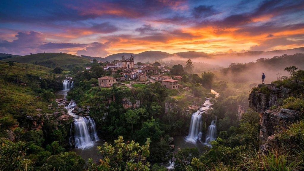 Cidade histórica cercada por cachoeiras e vegetação, com igreja, cânions e céu colorido ao pôr do sol.