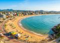Vista aérea de praia urbana em baía tranquila, com faixa de areia, mar azul, quiosques, ciclovia e cidade cercada por morros ao fundo.