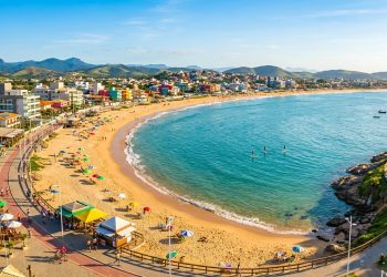 Vista aérea de praia urbana em baía tranquila, com faixa de areia, mar azul, quiosques, ciclovia e cidade cercada por morros ao fundo.