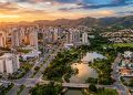 Vista aérea de cidade com prédios altos, avenidas, parque com lagos e montanhas ao fundo durante o pôr do sol.