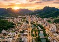 Vista aérea de cidade entre montanhas verdes ao pôr do sol, com prédios, avenidas iluminadas e rio atravessando a área urbana.