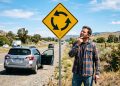 Homem confuso observa uma placa amarela de rotatória ao lado de uma rodovia, com carro parado no acostamento.