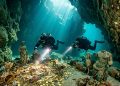 Mergulhadores exploram uma caverna submersa com moedas de ouro, ânforas, estátuas antigas e feixes de luz atravessando a água.