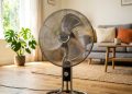 Ventilador ligado em uma sala de estar iluminada, com sofá, mesa de centro, planta e janela ao fundo.