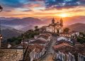 Cidade histórica com igreja colonial e casas antigas em rua de pedra, cercada por montanhas ao pôr do sol.