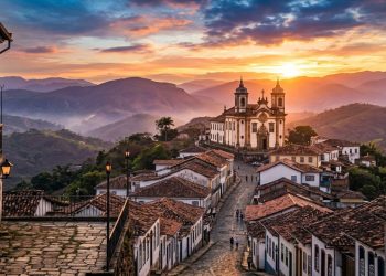 Cidade histórica com igreja colonial e casas antigas em rua de pedra, cercada por montanhas ao pôr do sol.