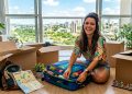 Mulher sorridente arruma mala colorida no chão de um apartamento, cercada por caixas de mudança, mapa de Sorocaba e vista urbana pela janela.