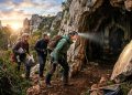 Equipe de exploradores com capacetes e lanternas observa a entrada de uma caverna rochosa ao entardecer, em paisagem natural montanhosa.