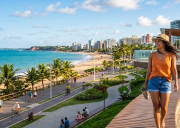Mulher caminha sorrindo por um calçadão elevado à beira-mar, com praia, ciclovia, coqueiros e prédios ao fundo.