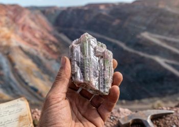 Mão segura um cristal bruto diante de uma mina a céu aberto, com bancadas de extração e anotações de campo ao fundo.