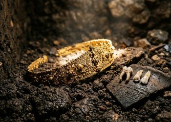 Tiara dourada ornamentada e pequenos dentes encontrados entre terra e pedras em uma escavação arqueológica.