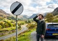 Homem parado ao lado de um carro em uma estrada rural, observando uma placa de fim de restrição em meio a montanhas verdes.