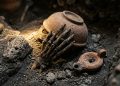 Fragmentos de cerâmica antiga e possível osso de mão parcialmente expostos em solo escuro durante escavação arqueológica.
