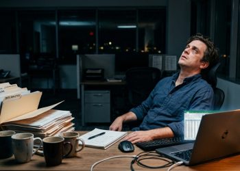 Homem sentado sozinho em escritório à noite, cercado por papéis, xícaras de café e notebook, com expressão de cansaço.