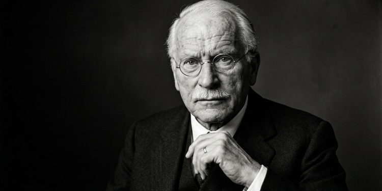 Homem idoso de óculos aparece em retrato preto e branco, com expressão séria e mão apoiada sob o queixo.