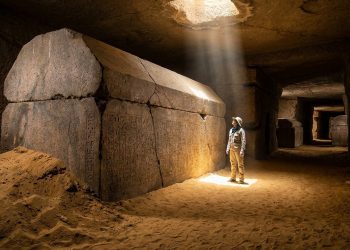 Pesquisador observa um grande sarcófago de pedra com inscrições antigas em uma câmara subterrânea iluminada por feixe de luz.