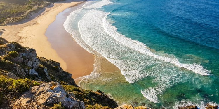 Ondas do mar curvam e chegam paralelas à praia ensolarada