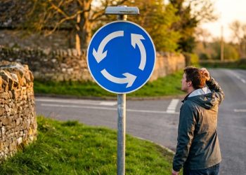 Homem observa uma placa azul de rotatória em uma estrada rural, com muro de pedra, casas antigas e vegetação ao redor.