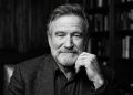 Retrato em preto e branco do ator Robin Williams com barba, expressão serena e reflexiva, com a mão no queixo e estantes de livros ao fundo.