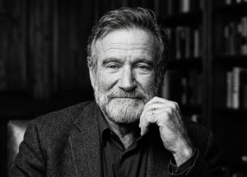 Retrato em preto e branco do ator Robin Williams com barba, expressão serena e reflexiva, com a mão no queixo e estantes de livros ao fundo.