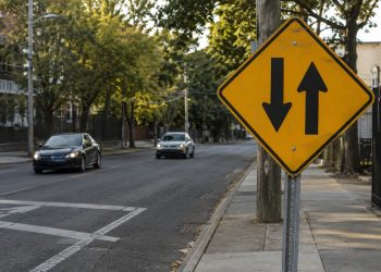 O que significa a placa de trânsito com duas setas pretas verticais paralelas apontando para sentidos opostos?