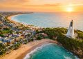 Vista aérea da orla de João Pessoa, com praias, mar azul, prédios urbanos e farol em área verde ao pôr do sol.