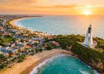 Vista aérea da orla de João Pessoa, com praias, mar azul, prédios urbanos e farol em área verde ao pôr do sol.