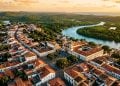 Vista aérea de centro histórico colonial com igreja, casarões antigos, telhados de barro e rio cercado por vegetação ao fundo.