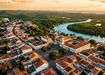 Vista aérea de centro histórico colonial com igreja, casarões antigos, telhados de barro e rio cercado por vegetação ao fundo.