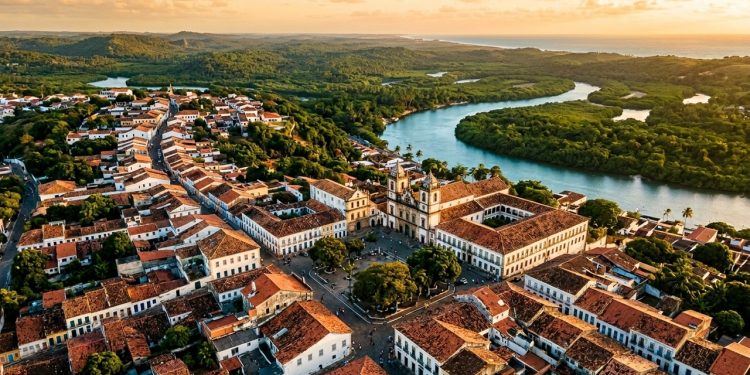Vista aérea de centro histórico colonial com igreja, casarões antigos, telhados de barro e rio cercado por vegetação ao fundo.