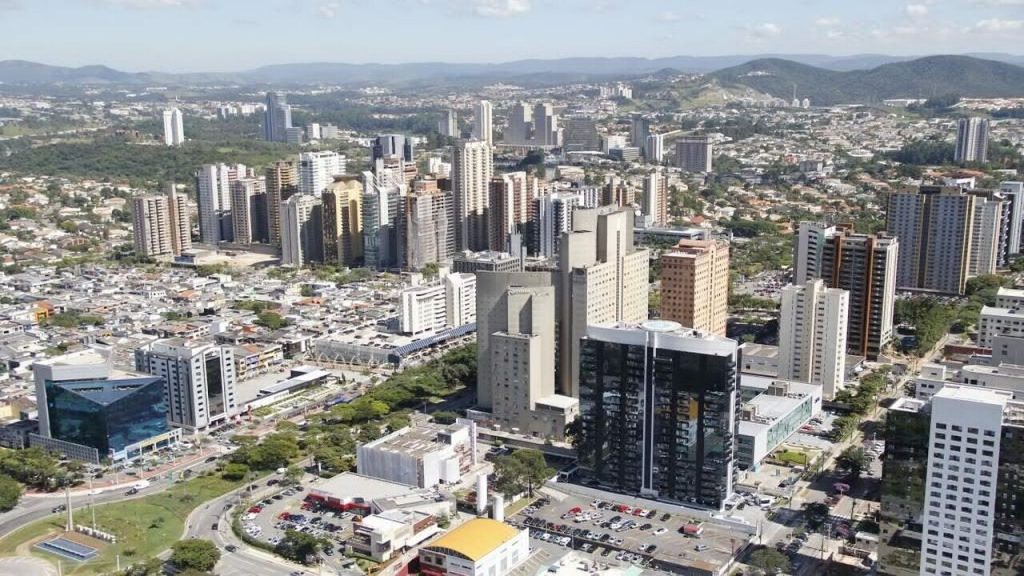 Vista aérea de Alphaville, em São Paulo, com prédios comerciais, áreas urbanas, avenidas e morros ao fundo.