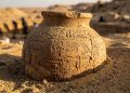 Vaso de cerâmica antigo com inscrições egípcias aparece parcialmente enterrado em área arqueológica no deserto.