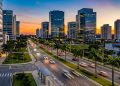 Avenida moderna em Barueri ao entardecer, com prédios comerciais, palmeiras, carros em movimento e céu colorido.