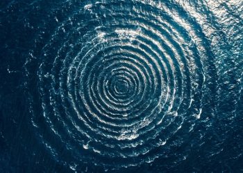 Ondas circulares se formando na superfície azul do oceano, vistas de cima, em padrão semelhante a um redemoinho.