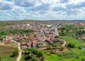 Vista aérea de drone da cidade de Cabaceiras, Paraíba, aninhada em um vale cercado por montanhas secas da Caatinga. As casas têm telhados de telhas vermelhas. Uma estrada de asfalto corta a cidade e os campos ao redor são verdes. O céu tem nuvens brancas e macias.