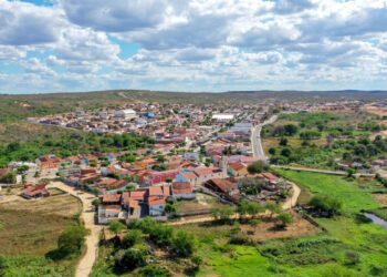 Vista aérea de drone da cidade de Cabaceiras, Paraíba, aninhada em um vale cercado por montanhas secas da Caatinga. As casas têm telhados de telhas vermelhas. Uma estrada de asfalto corta a cidade e os campos ao redor são verdes. O céu tem nuvens brancas e macias.