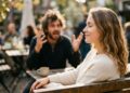 Fotografia diurna em um ambiente de café ao ar livre. Em primeiro plano e em foco nítido, uma mulher sentada apresenta uma expressão de total serenidade, com os olhos fechados e um leve sorriso, ignorando o que acontece atrás dela. Ao fundo, totalmente desfocado, um homem aparece gesticulando de forma agressiva e irritada.