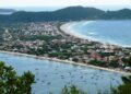 Vista panorâmica do alto de um morro revelando a geografia estreita da península de Bombinhas. À esquerda, as águas calmas da Baía de Canto Grande, pontilhadas por dezenas de pequenos barcos de pesca. À direita, a longa faixa de areia e as ondas do mar aberto da Praia de Mariscal. No meio, a cidade construída sobre a estreita faixa de terra cercada por morros verdes.