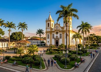 Praça central arborizada com igreja matriz histórica, palmeiras, jardins, coreto, pessoas caminhando e rua de pedra ao entardecer.