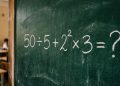 Quadro verde em sala de aula com a expressão matemática 50 ÷ 5 + 2² × 3 = ? escrita em giz branco.