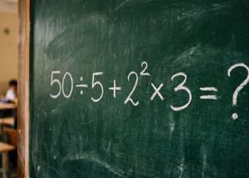 Quadro verde em sala de aula com a expressão matemática 50 ÷ 5 + 2² × 3 = ? escrita em giz branco.