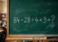 Quadro verde em sala de aula com a expressão matemática 84 - 28 ÷ 4 × 3 = ? escrita em giz branco.