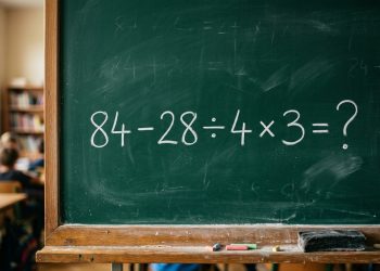 Quadro verde em sala de aula com a expressão matemática 84 - 28 ÷ 4 × 3 = ? escrita em giz branco.