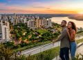 Casal observa cidade do Oeste do Paraná ao pôr do sol, com prédios, áreas verdes, rio e mirante panorâmico.