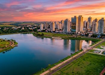 Vista panorâmica de cidade brasileira ao pôr do sol, com lago urbano, prédios, áreas verdes e ciclovia ao redor.